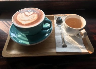 Üstünde kalp latte sanatı olan bir fincan kahve. Kağıt mendilin üzerinde kahve fincanının yanında bir fincan çay var. Her şey ahşap tepside. Onlar için yumuşak bir güneş ışığı var..