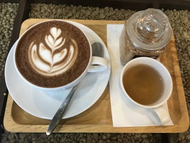 Üstünde latte sanatının olduğu sıcak kahve. Kağıt mendilin üzerinde kahve fincanının yanında bir fincan çay var. Her şey ahşap tepside. Arka plan fındıklı kahve..