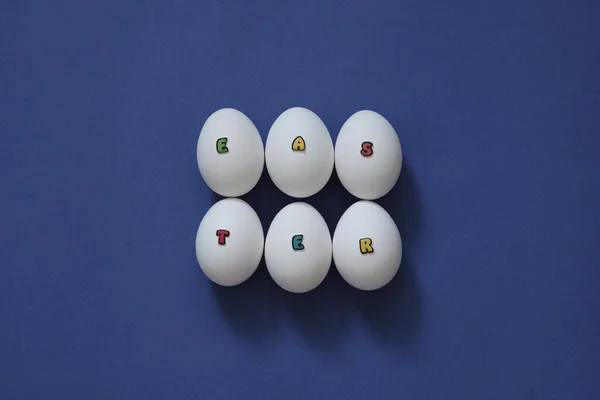 Творче компонування білих курячих яєць з написом Великдень з кольорових літер на синьому фоні . — стокове фото