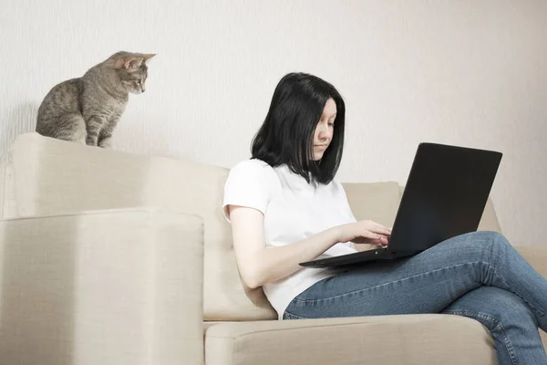 Het meisje gebruikt een laptop voor werk op afstand. Een grijze kat zit op de bank en volgt zorgvuldig het werk van de freelancer. — Stockfoto