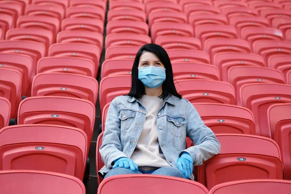 A female fan in a medical mask and rubber gloves sits alone in an empty stadium with red seats. Cancellation of sporting events during the coronavirus. One man army concept.