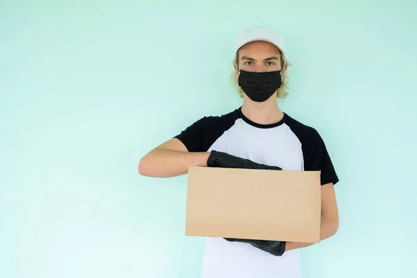 Siyah kısa kollu, beyaz şapkalı, siyah koruyucu maskeli beyaz tişörtlü bir adam elinde bir karton kutu tutuyor. Karantina sırasında eve teslim