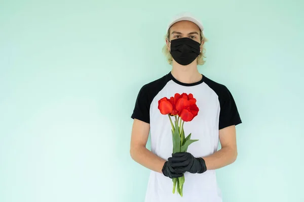a guy in a white T-shirt with a black short sleeve and a white cap, in a black protective mask, is holding a bouquet of red tulip flowers in his hands Home delivery during quarantine