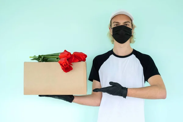Siyah kısa kollu, beyaz şapkalı, siyah koruyucu maskeli beyaz tişörtlü bir adam kırmızı lalelerin olduğu karton bir kutu tutuyor. Karantina sırasında eve teslim