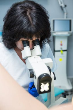 Siyah saçlı, beyaz önlüklü ve mavi steril eldivenli bir jinekolog mikroskobu olan bir hastayı muayene ediyor. Üreme tıbbı