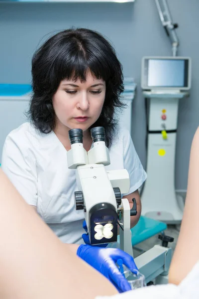 Siyah saçlı, beyaz önlüklü ve mavi eldivenli bir jinekolog mikroskobu olan bir hastayı muayene ediyor.