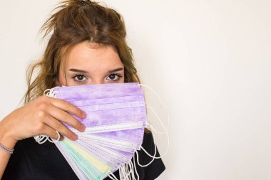 Güzel kahverengi gözlü kız elinde bir sürü renkli hijyenik maske tutuyor, burnunu ve ağzını kapatıyor. Değişik maskeler. Seçimini yap. Maske koleksiyonu. Yatay yerleşim planı. Boşluğu kopyala