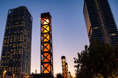 İki gökdelen ve arkaplanda mavi günbatımı gökyüzü olan bir sıra yıldırım aydınlatma sütunu. Gün batımından sonra şehir manzarası. Şehir hayatı. Barselona İspanya 'da ofis siluetleri inşa ediliyor