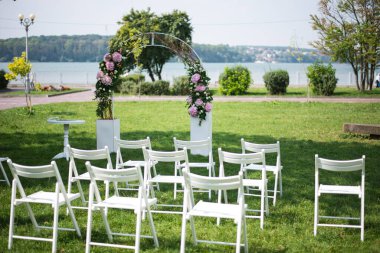 Pembe taze kesilmiş ortanca ve kremalı güllerle süslenmiş güzel bir düğün kemeri ve arka planda gölü olan konuklar için sandalyeler.