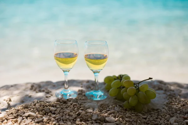 Wine glasses filled with white semi-sweet tokaj wine and bunch of green grapes located at the flat stone on pebble beach. Sea at the background. Horizontal view. Close-up