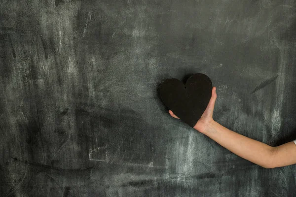 Black wooden Heart in Hand of a Kid in chalkboard background. Black Heart. Place for text on Left side of image. Chalkboard texture. Good-bye school concept. Horizontal layout copy space.