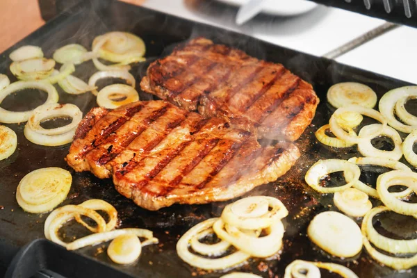 两片猪排牛排和洋葱切成片，在电炉上烤着，在烤过的食物上可以看到烟熏的痕迹 — 图库照片