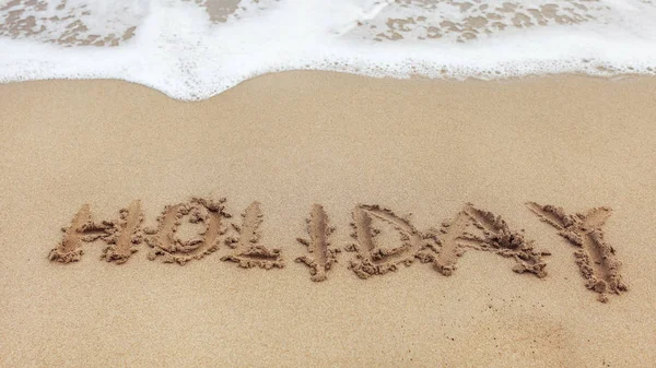 Word HOLIDAY written on wet sand of beach, small spot of white s — Stock Photo, Image