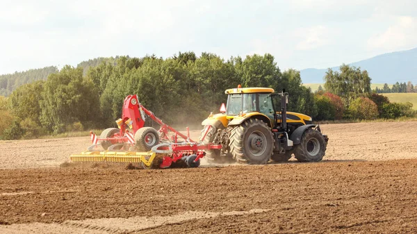 Жовтий трактор тягне червоний механізм сівби над полем, дерева позаду. — стокове фото