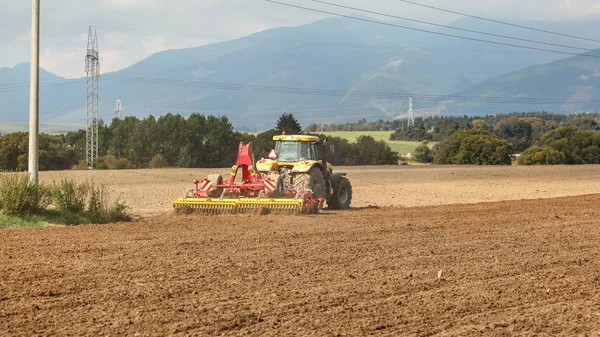 Sárga traktor húzza piros vetési mechanizmus felett száraz területen, fák — Stock Fotó