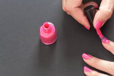 Pembe tırnak cilası sürülüyor. Kadın eli, küçük fırça, cila şişesi ve gri arka planda tırnakların yakın çekimi.