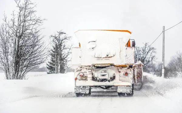 Orange plough truck on snow covered road; gray sky and trees in background; view from car driving behind - winter road maintenance