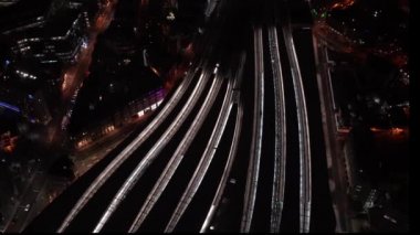 Tren istasyonunun gece görüşü, platformlardan yavaş hareket eden trenler, caddelerde giden arabalar, platformlarda yürüyen yayalar.