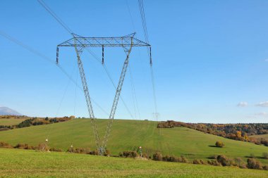 Kırsal kesimlerde yeşil otlarla kaplı tepeler ve küçük ormanlarla inşa edilmiş büyük metal elektrik direği, mavi gökyüzü arka planı.
