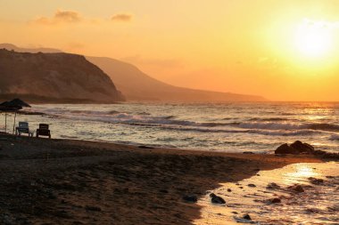 Gün batımı boş bir kumsalda iki güneşli yatağa ışıldar. Küçük deniz dalgaları, sisli arka plandaki tepeler