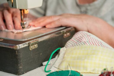 Eski dikiş makinesi üzerinde çalışan yaşlı bir kadın. Coronavirus 'a karşı yüz maskesi yapıyor. Yayılıyor, ön plandaki bitmiş maskelere odaklanıyor.