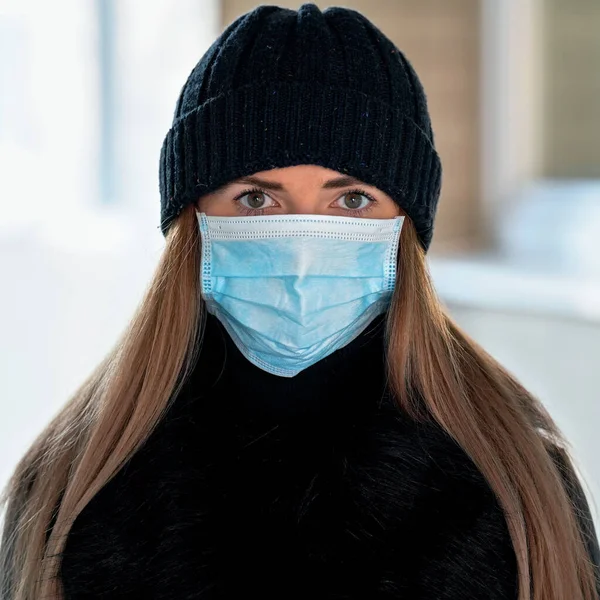 Young woman wearing blue disposable virus face mouth nose mask and warm beanie hat, closeup portrait, blurred background. Coronavirus covid-19 outbreak prevention illustration