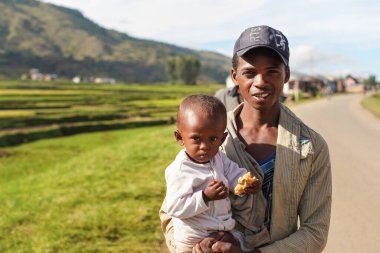 Manandoana, Madagaskar - 26 Nisan 2019: Bilinmeyen Malagasy adamı elinde yiyecek parçasıyla çocuğu tutuyor, güneşli bir günde arka planda pirinç tarlaları. Afrika 'nın bu bölgesindeki insanlar fakir ama neşelidir.