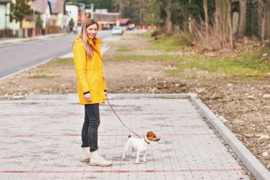 Sarı ceketli, gülümseyen ve kameraya bakan genç bir kadın. Pembe tasmalı teriyer köpeği, arka planda bulanık evler.