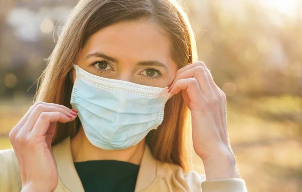 Young woman wearing blue disposable virus mouth nose mask, touching it with hands, nice sunny bokeh in background, closeup face portrait