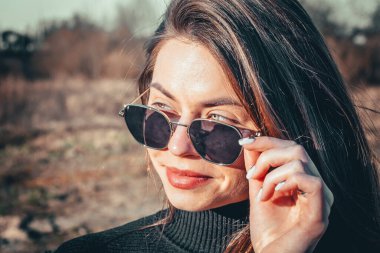 Esmer bir kız açık havada siyah kürk ceket ve siyah güneş gözlüğü giyerek yürüyor..