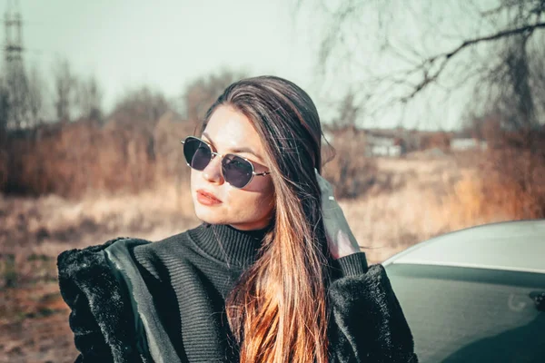 Esmer bir kız açık havada siyah kürk ceket ve siyah güneş gözlüğü giyerek yürüyor..