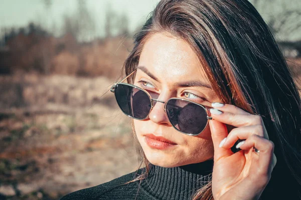 Esmer bir kız açık havada siyah kürk ceket ve siyah güneş gözlüğü giyerek yürüyor..