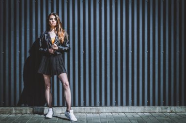 Siyah arka planda parlak makyajlı bir güzellik var. O, siyah bir ceket ve kısa bir etek giydi, külotlu çorabı üzerinde bir ağ ve beyaz spor ayakkabıları vardı..