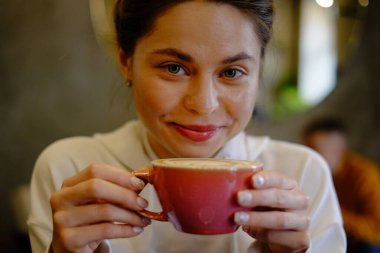 Bir kafede kapuçino içen güzel genç bir kız kalp şeklinde kahve çiziyor, pembe kupa, kahve içiyor ve eğleniyor.