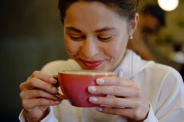 Bir kafede kapuçino içen güzel genç bir kız kalp şeklinde kahve çiziyor, pembe kupa, kahve içiyor ve eğleniyor.