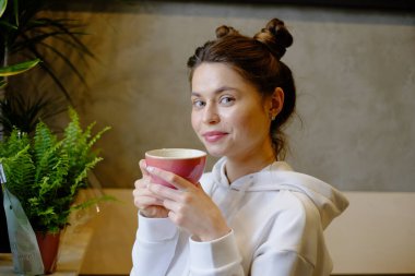 Bir kafede kapuçino içen güzel genç bir kız kalp şeklinde kahve çiziyor, pembe kupa, kahve içiyor ve eğleniyor.