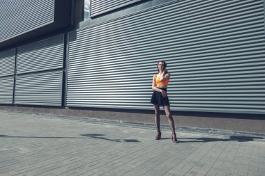 Siyah bir binanın arka planında büyüyen genç ve güzel bir esmer doğrudan kameraya bakıyor, parlak turuncu bir üst, siyah balıkçı ağı taytı ve siyah deri bir ceket giyiyor.