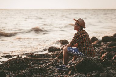 Bir adam yazın gün batımında ve deniz suyunun yanındaki taşın üzerinde oturuyor..