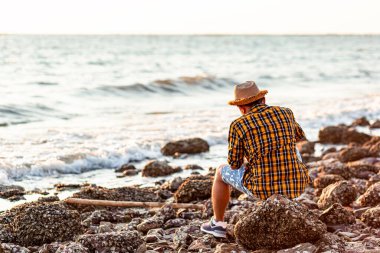 Bir adam yazın gün batımında ve deniz suyunun yanındaki taşın üzerinde oturuyor..