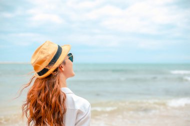 Yaz tatilinde şapkalı, elbiseli ve güneş gözlüklü mutlu kadın adanın kumsalındaki manzaranın tadını çıkarıyor..