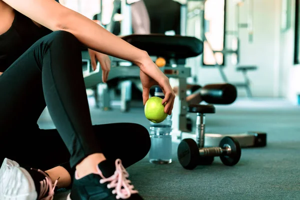Spor salonundaki kadın egzersizi ara verip rahatlıyor. Spor ve dambıl eğitiminden sonra elmanın meyvesini tutarken yerde su şişesi..