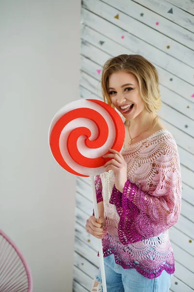 Aantrekkelijke blonde meisje verblijf in helder roze gebreide trui en eten grote rode witte lollipop — Stockfoto