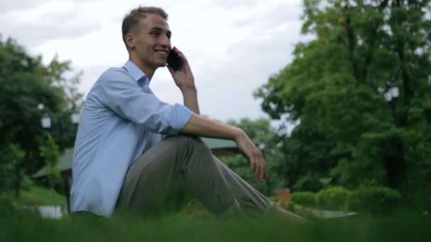 Парень сидит на траве и говорит по телефону против зеленых растений — стоковое видео