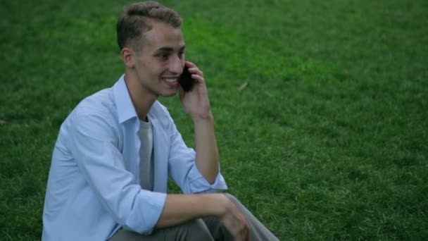 Chico se sienta en la hierba y tiene conversación en el teléfono en el parque — Vídeos de Stock