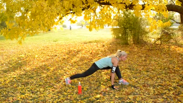 有吸引力的运动金发女郎在公共公园的地面上进行分裂和锻炼 上面覆盖着一只黄色的叶子 — 图库视频影像