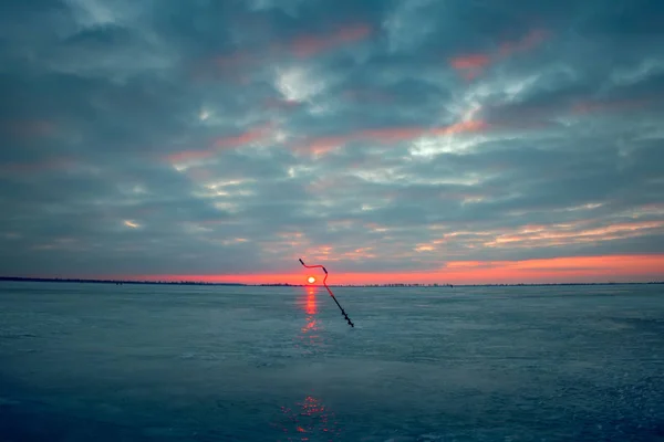Furadeira de gelo no fundo do pôr do sol em um lago de inverno . — Fotografia de Stock