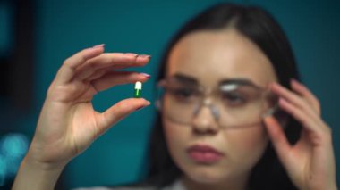 Young attractive female scientist with protective eyeglasses holding a transparent pill with fingers in gloves in the pharmaceutical research laboratory