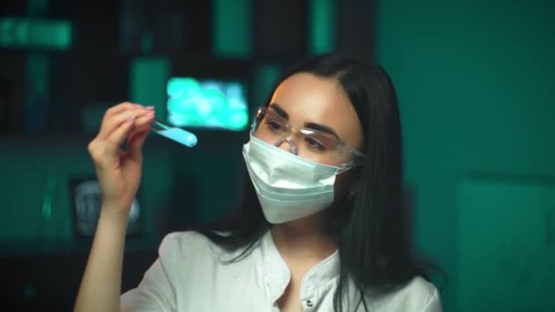 Uma assistente de laboratório com óculos de proteção e uma máscara médica segura um frasco de solução azul na frente dela e olha para ele — Vídeo de Stock