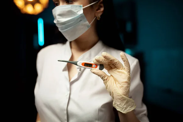 Maskeli güzel genç kadın doktor portresi ve siyah arka planda elektronik termometre tutan tıbbi eldivenler..