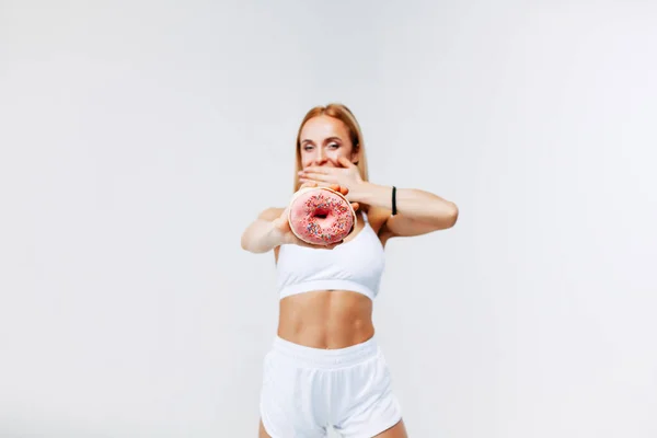 Spor elbiseli sıska bir kadın, elleri ile ağzını kapatıyor. Hata yapmaktan utanıyor, korkunun dışa vurumu, sessizlikten korkuyor ve beyaz bir arka planda donut tutuyor.